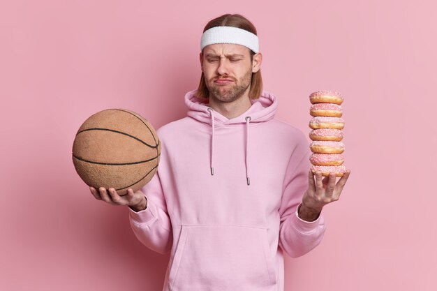 La gente se divierte el concepto de comida chatarra. El hombre deportivo disgustado mira con tristeza la pelota de baloncesto tiene un montón de dulces y apetitosos donuts no quiere jugar vestido con una sudadera con capucha.