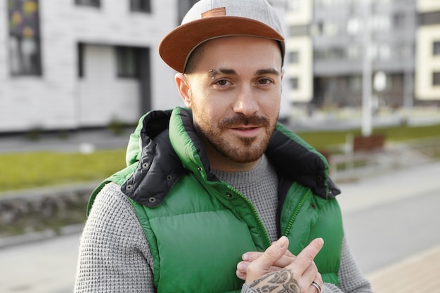 Gente, estilo de vida moderno, estilo urbano y concepto de moda callejera. Chico joven sin afeitar con estilo seguro con ceja perforada y tatuaje en la mano con un gran plan, caminando al aire libre en las calles de la ciudad