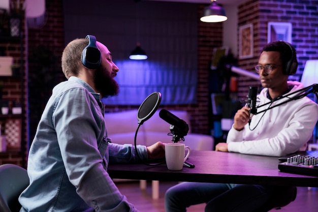 Gente feliz grabando podcasts en medios sociales con equipos de blogs, usando la estación vlog para transmitir conversaciones en vivo en el canal de Internet. Hombres reunidos en el estudio para grabar un programa de entrevistas.