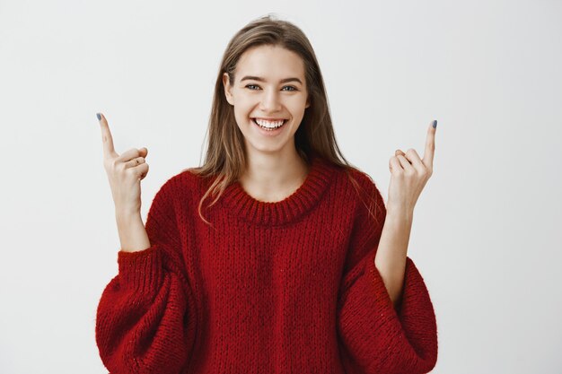 La gente necesita levantar la cabeza y vivir del lado positivo. Hermosa estudiante satisfecha con suéter holgado de moda, levantando los dedos índices y apuntando hacia arriba con una amplia sonrisa de satisfacción sobre la pared gris