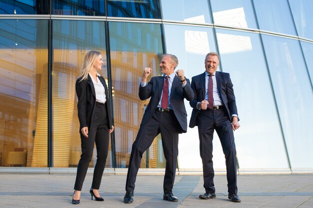 Gente de negocios alegre celebrando la victoria, de pie juntos en la fachada del edificio de oficinas de cristal. Vista frontal de cuerpo entero. Equipo exitoso y concepto de trabajo en equipo