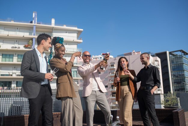 Gente de negocios alegre en fiesta en la azotea de la terraza. Hombres y mujeres con ropa formal de pie en el techo de la terraza, abriendo champán, riendo. Teambuilding, concepto de fiesta