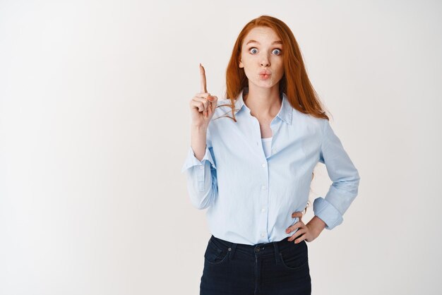 Gente de negocios Atractiva gerente femenina con cabello pelirrojo y ojos azules, labios fruncidos levantando el dedo y apuntando hacia arriba mostrando el fondo blanco del anuncio