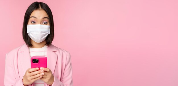 Gente de negocios y concepto covid19 Joven empresaria asiática en traje y mascarilla médica usando una aplicación de teléfono inteligente de pie sobre fondo rosa