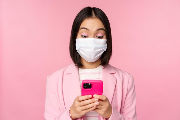 Gente de negocios y concepto covid19 Joven empresaria asiática en traje y mascarilla médica usando una aplicación de teléfono inteligente de pie sobre fondo rosa