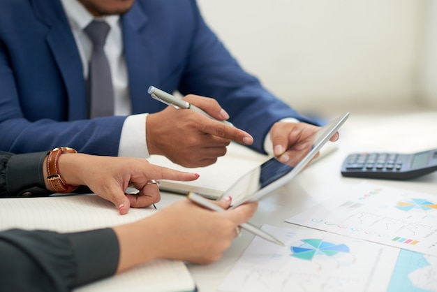 Gente de negocios irreconocible sentada en una reunión con gráficos, mirando y señalando la tableta