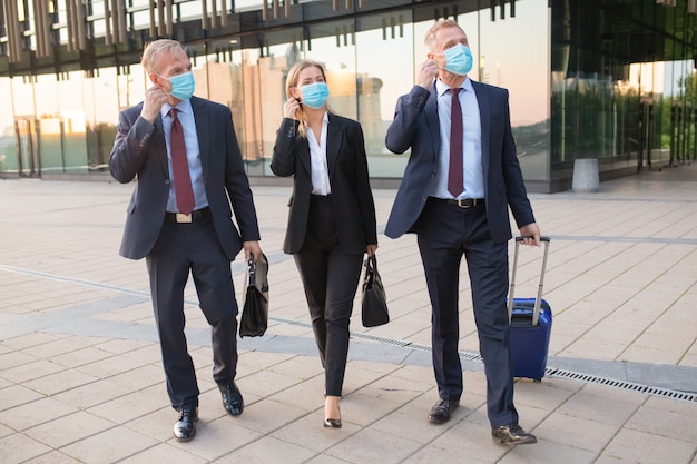 Gente de negocios que se ajusta o está lista para quitarse las máscaras faciales mientras camina con el equipaje al aire libre, cerca de los edificios de oficinas. Viaje de negocios y fin del concepto de epidemia.