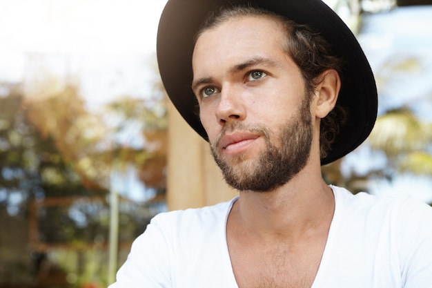 Gente y ocio. Disparo en la cabeza del hombre joven de moda con barba de moda con sombrero negro con expresión pensativa, mirando a la distancia, día de planificación