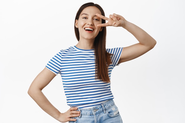 Gente positiva y brillante La niña riendo feliz muestra un signo de paz cerca de los ojos con un aspecto alegre de pie en una camiseta de verano con un fondo blanco estampado a rayas