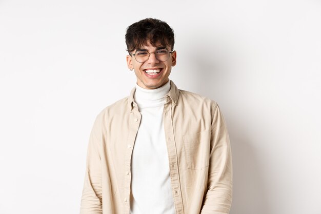 Gente real. Retrato natural de chico feliz sonriendo y riendo, mirando optimista a la cámara, de pie con gafas sobre fondo blanco.