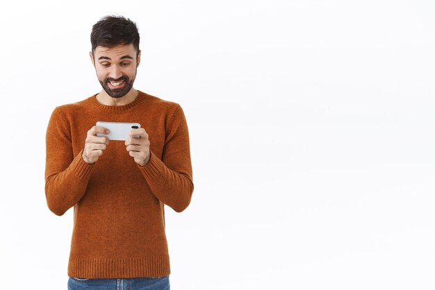 Gente de tecnología y concepto de comunicación Retrato de hombre barbudo caucásico feliz regocijándose sonriendo en el teléfono móvil mientras juega una aplicación de juego impresionante disfrutando de carreras o arcade fondo blanco