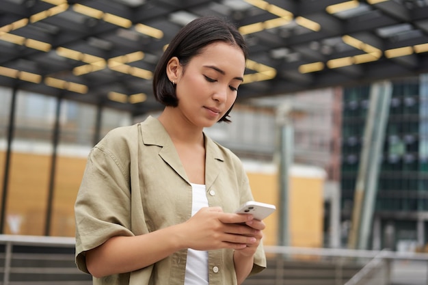 Gente de tecnología joven sonriente chica asiática sosteniendo teléfono inteligente usando la aplicación móvil mientras está de pie en str