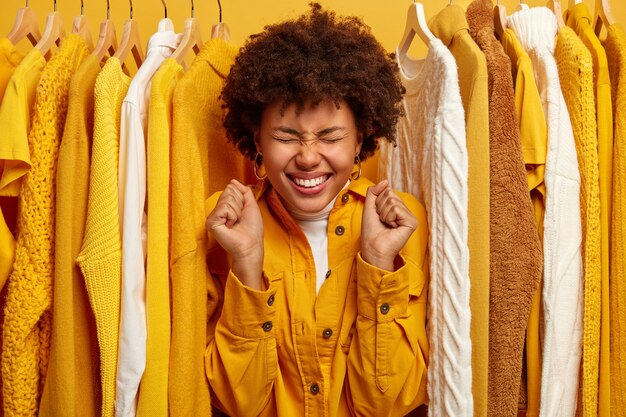 Gente, vestirse, concepto de compras. Alegre mujer de piel oscura con una gran sonrisa, aprieta los puños, se encuentra en el armario entre la ropa