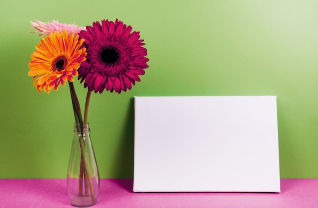 Gerbera florece en el florero cerca de la tarjeta en blanco en el escritorio rosado contra la pared verde