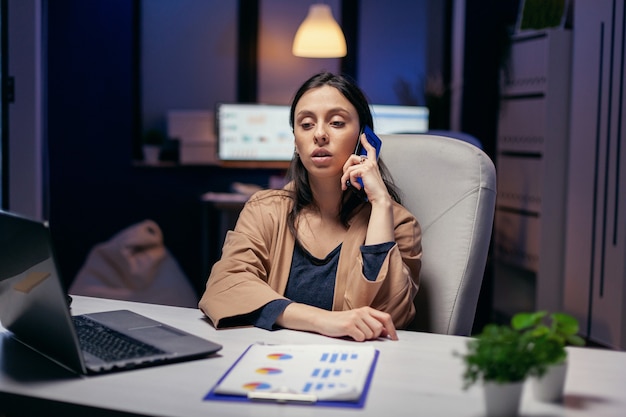 Gerente adicto al trabajo hablando con el cliente por teléfono por la noche. Mujer emprendedora que trabaja hasta altas horas de la noche en negocios corporativos haciendo horas extraordinarias en el curso de una llamada telefónica.