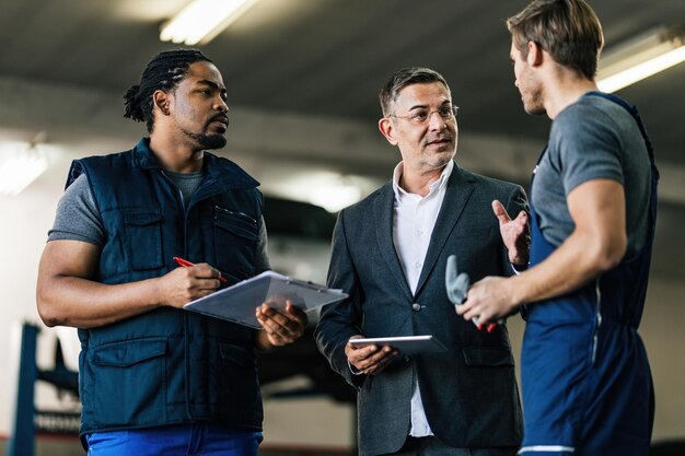 Gerente adulto medio que usa una tableta digital mientras habla con mecánicos de automóviles en un taller de reparación de automóviles