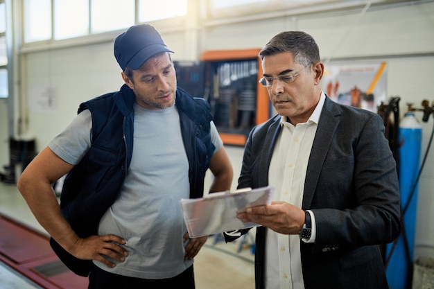 Gerente de adultos medianos y mecánico de automóviles analizando datos mientras revisa informes en el taller de reparación de automóviles