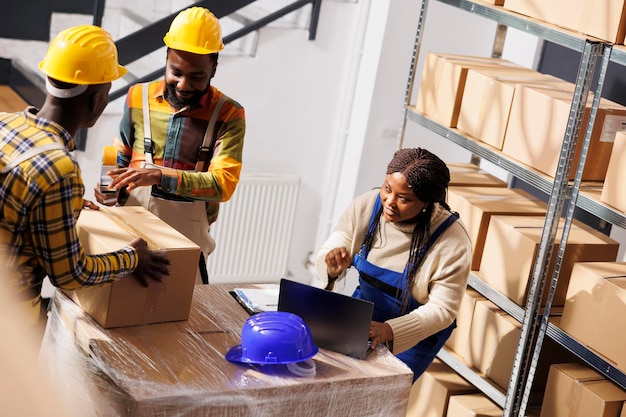 Foto gratuita gerente de almacén revisando la lista de pedidos y coordinando el proceso de empaque de paquetes. supervisora afroamericana con computadora portátil que controla el sellado y envío de cajas de cartón de carga