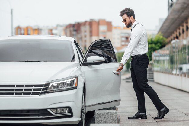 Gerente barbudo guapo entrando en su automóvil mientras está parado al aire libre en las calles de la ciudad cerca del moderno centro de oficinas