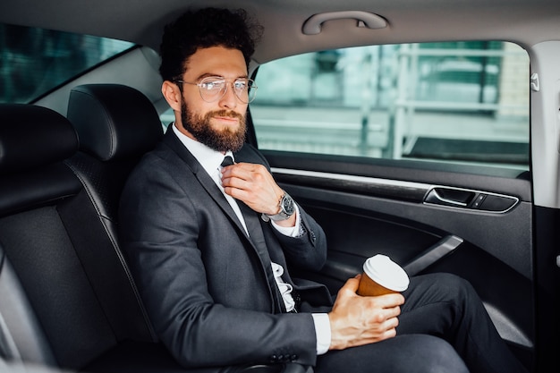 Gerente barbudo guapo sentado con café para ir en el asiento trasero del auto nuevo