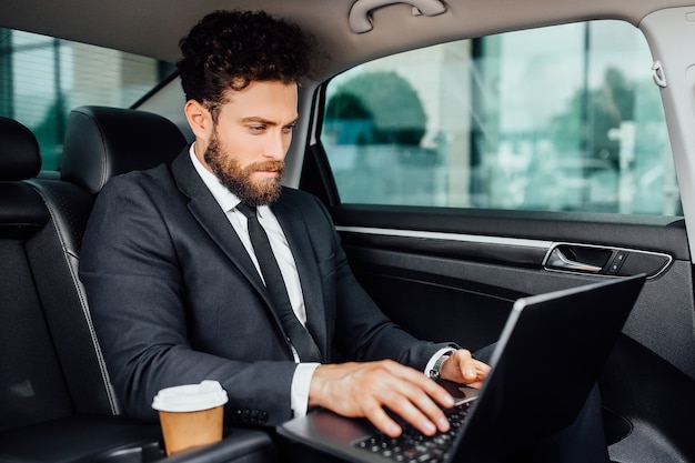 Gerente barbudo guapo trabajando en su computadora portátil con café para ir en el asiento trasero del auto nuevo