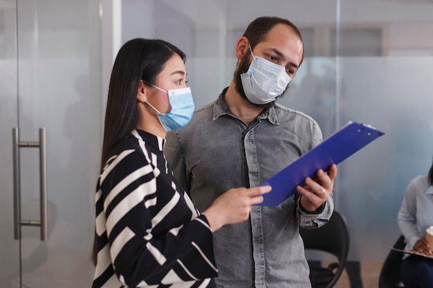 Foto gratuita el gerente del departamento de recursos humanos revisa el currículum vitae del solicitante de empleo en el portapapeles mientras se encuentra en el área del vestíbulo de la empresa de nueva creación. el empleador y el futuro empleado usan una máscara protectora mientras están en la sala de espera.