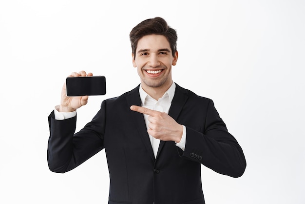 Gerente de director general de trabajador de oficina masculino confiado que señala la pantalla horizontal del teléfono y una sonrisa demuestra el sitio web o la aplicación de pie en traje contra el fondo blanco