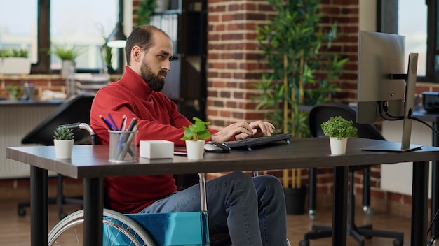 Foto gratuita gerente con discapacidad que trabaja en el crecimiento del negocio con una computadora en el escritorio, en silla de ruedas. trabajador masculino que planifica una estrategia financiera para crear un desarrollo ejecutivo de la empresa.