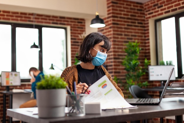 Gerente ejecutivo con mascarilla de protección médica para prevenir la infección con covid19 analizando gráficos comerciales escribiendo estrategia de gestión en computadora portátil. Empresaria trabajando en ideas de proyectos