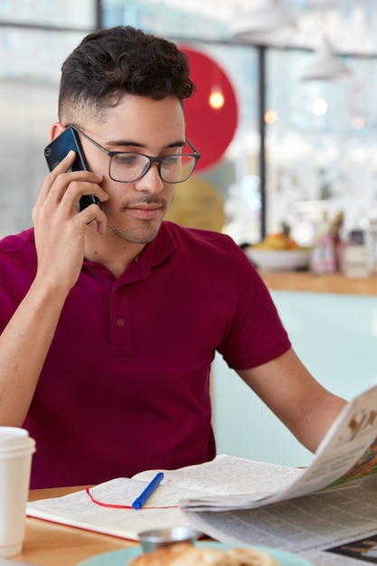 Gerente ejecutivo masculino serio confiado lee noticias financieras en el periódico diario, tiene conversación telefónica, modelos en la mesa en la cafetería con bebida fresca, toma notas en el bloc de notas. Tiro vertical