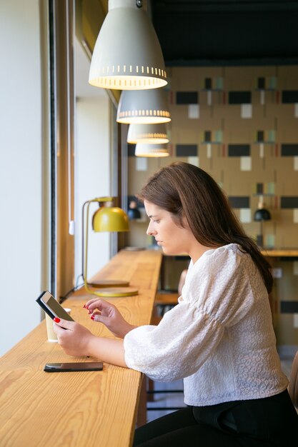 Gerente femenina enfocada usando tableta mientras está sentado en el escritorio en el espacio de trabajo conjunto o en la cafetería