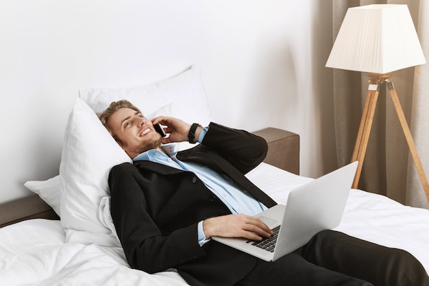 Gerente guapo y feliz con elegante corte de pelo y barba acostado en la cama en la habitación del hotel, hablando por teléfono y revisando su trabajo en la computadora portátil antes de la reunión de negocios