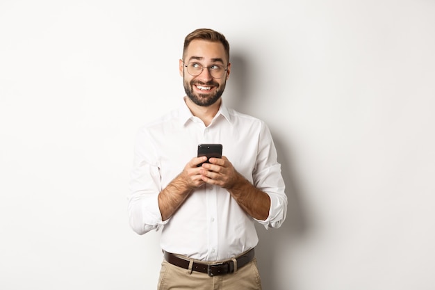 Foto gratuita gerente guapo joven que usa el teléfono móvil y piensa en contestar el mensaje, mirando la esquina superior izquierda y sonriendo, de pie