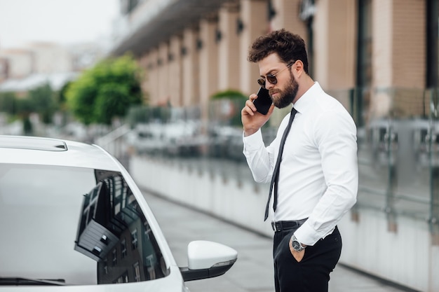 Gerente guapo, serio y barbudo con gafas de sol, hablando por teléfono y parado cerca de su automóvil al aire libre en las calles de la ciudad cerca del moderno centro de oficinas