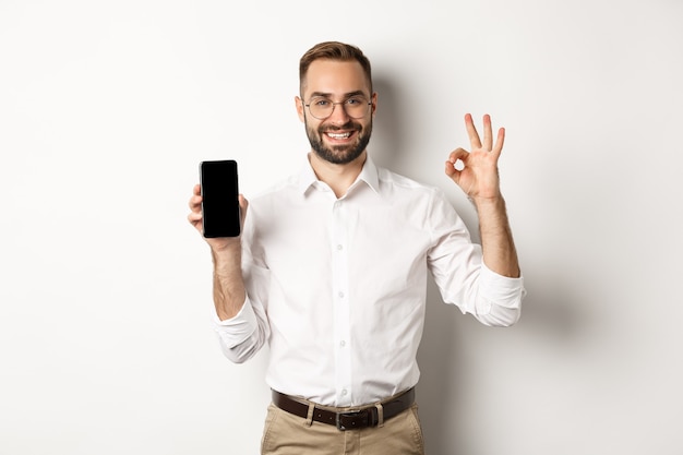 Gerente joven satisfecho que muestra la pantalla del teléfono inteligente y el signo bien, recomendando la aplicación, de pie sobre fondo blanco.