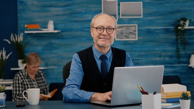 Gerente jubilado sentado en el escritorio frente a la cámara sonriendo después de escribir en la computadora portátil trabajando desde casa mientras la esposa mayor lee un libro en segundo plano.