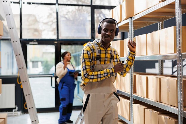 Gerente de logística afroamericano bailando en el almacén de la fábrica y mirando la cámara. Joven vistiendo ropa de trabajo escuchando música y haciendo movimientos de manos en el almacén