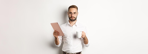 Foto gratuita gerente masculino confiado leyendo trabajo en tableta digital y bebiendo café de pie sobre backgr blanco