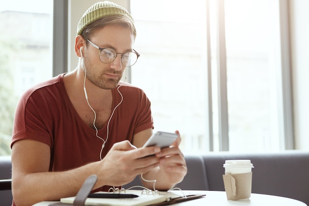 Gerente masculino profesional escucha música en línea en auriculares blancos, se sienta en la mesa rodeado con su diario