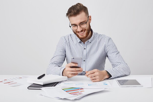 Gerente masculino sonriente rodeado de muchos papeles y gadgets, recibe felicitaciones por teléfono celular de un amigo como cumpleaños