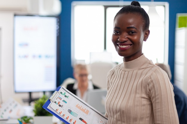 Gerente de mujer africana mirando a la cámara sonriendo, sosteniendo el portapapeles, mientras diversos compañeros de trabajo hablando en segundo plano.