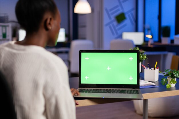 Gerente de mujer africana trabajando en una computadora portátil con maqueta de pantalla verde, escritorio con clave de croma sentado en el escritorio en la oficina de negocios tarde en la noche