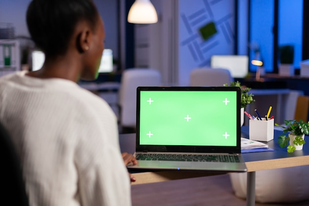 Gerente de mujer africana trabajando en una computadora portátil con maqueta de pantalla verde, escritorio con clave de croma sentado en el escritorio en la oficina de negocios tarde en la noche