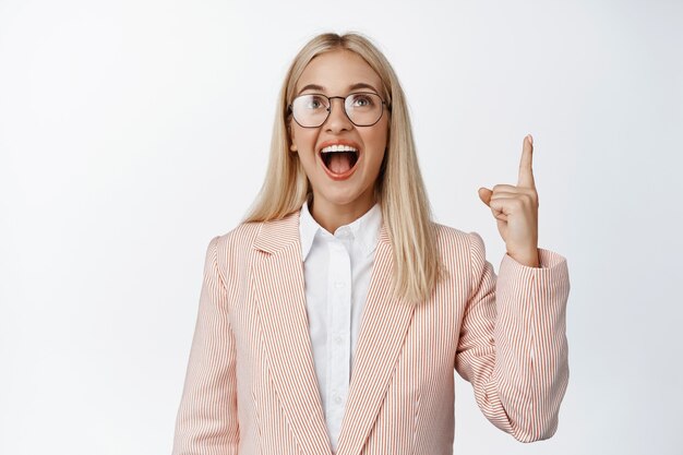 Gerente de mujer emocionada, empleada con gafas y traje apuntando y mirando hacia arriba con cara de asombro en blanco.