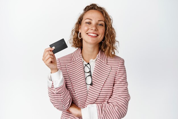 Foto gratuita gerente mujer feliz muestra tarjeta de crédito y sonríe complacida, se encuentra en traje en blanco