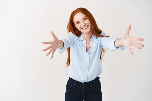 Gerente de mujer pelirroja feliz extendiendo las manos hacia adelante para abrazarte. Linda mujer con cabello pelirrojo estirando los brazos para abrazos, de pie sobre la pared blanca