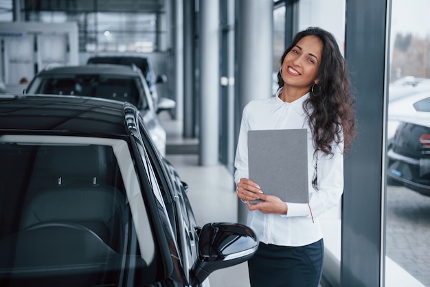 Gerente de mujer de pelo rizado se encuentra cerca del coche en el salón del automóvil