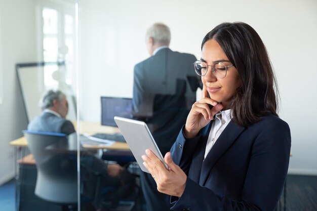 Gerente de mujer pensativa en gafas mirando en la pantalla de la tableta y sonriendo mientras dos empresarios maduros discutiendo el trabajo detrás de la pared de vidrio. Copie el espacio. Concepto de comunicación