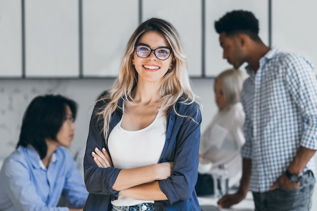 Gerente mujer rubia segura posando con una sonrisa después de la conferencia con otros empleados. Programador asiático hablando con autónomo africano mientras secretaria rubia riendo.