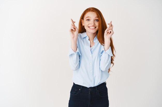 Gerente de mujer sonriente esperanzada con cabello rojo y ojos azules, pidiendo deseos, esperando resultados con los dedos cruzados para la buena suerte, pared blanca